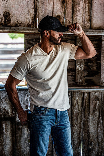 Man adjusting his cap while wearing a natural heather V-neck cotton jersey top and jeans near a barn window