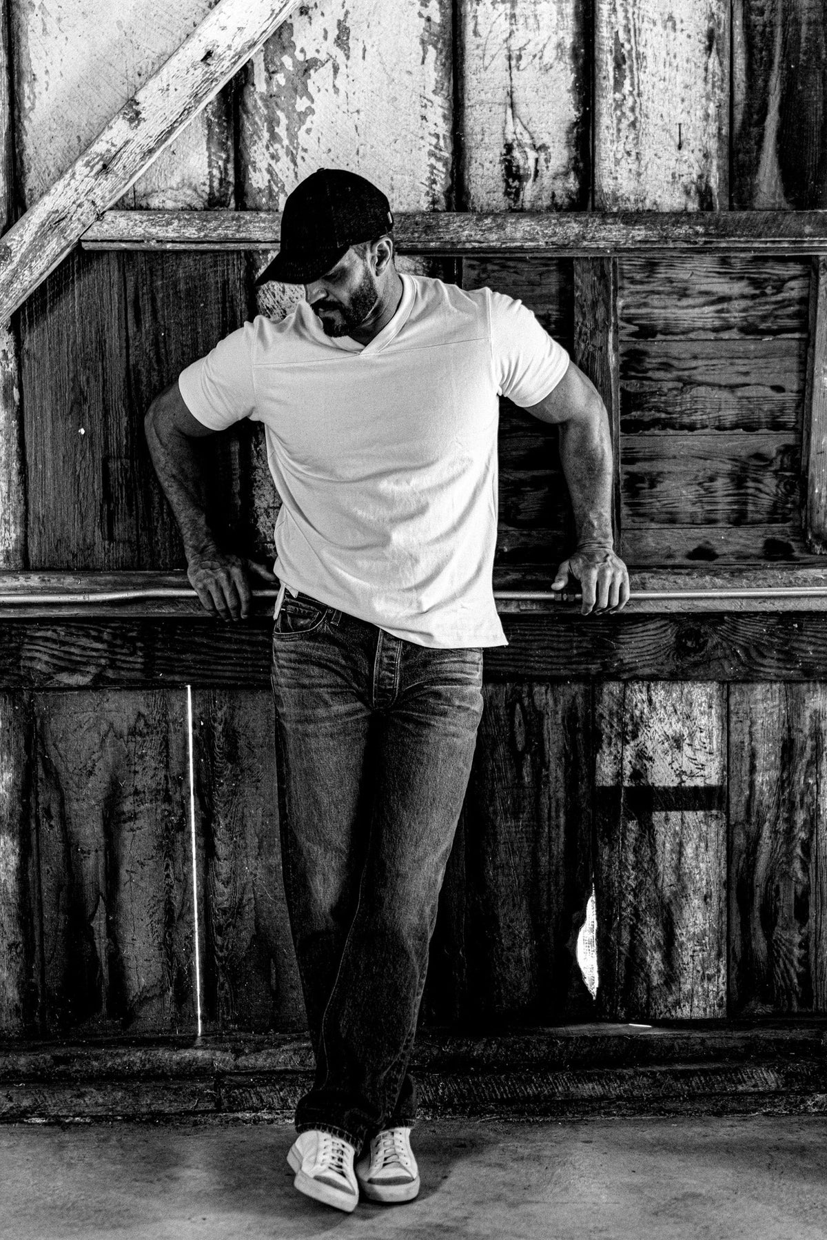 Black and white photo of a man in a men's V-neck cotton jersey top and jeans leaning against a railing in a wooden barn