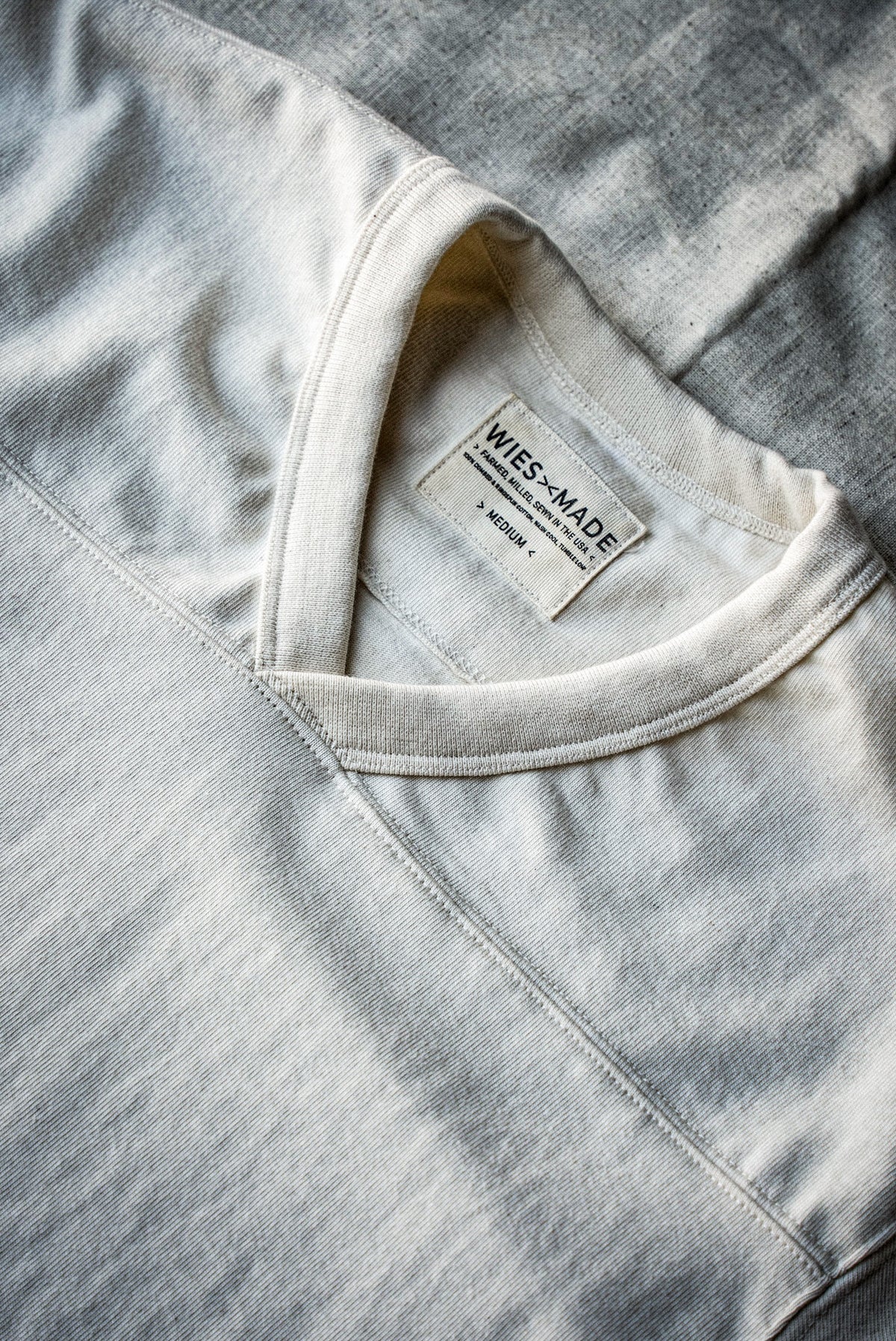 Flat-lay close-up of a men's natural heather V-neck cotton jersey top, showing the ribbed collar and interior size label
