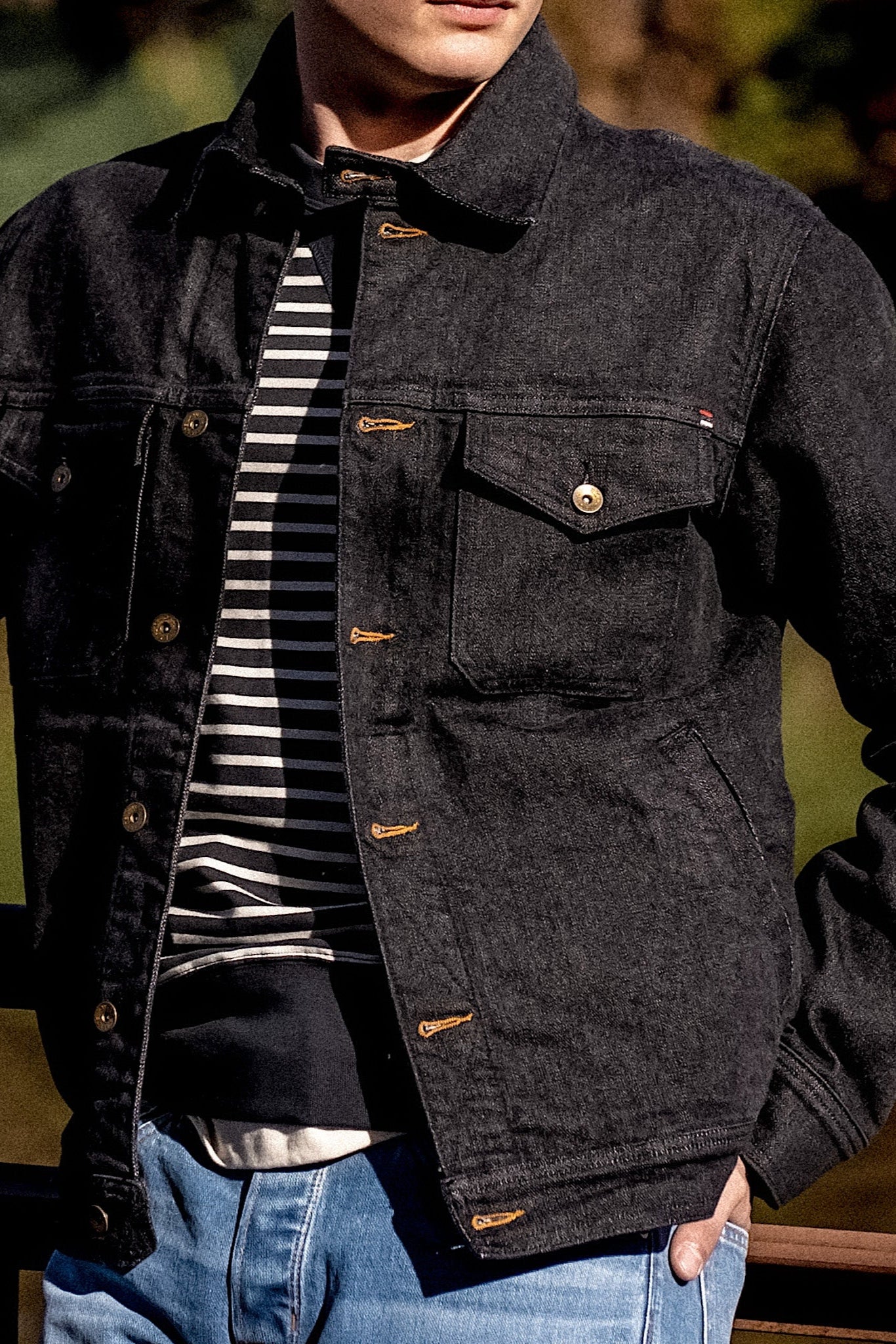 Dark denim jacket with button closures and chest pockets, layered over a striped sweater and paired with blue jeans in sunlight.