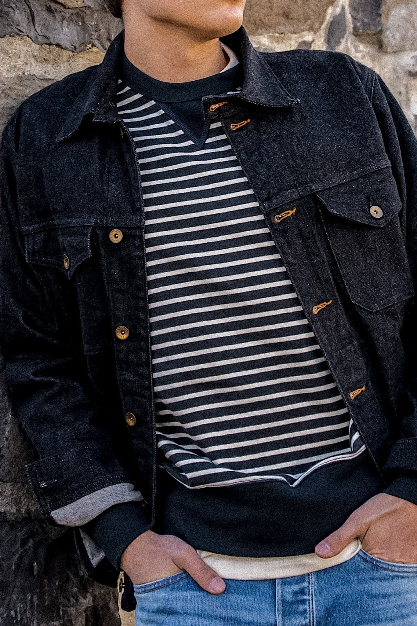 Real denim jacket styled with a striped sweater and jeans, worn by a person leaning against a stone wall.