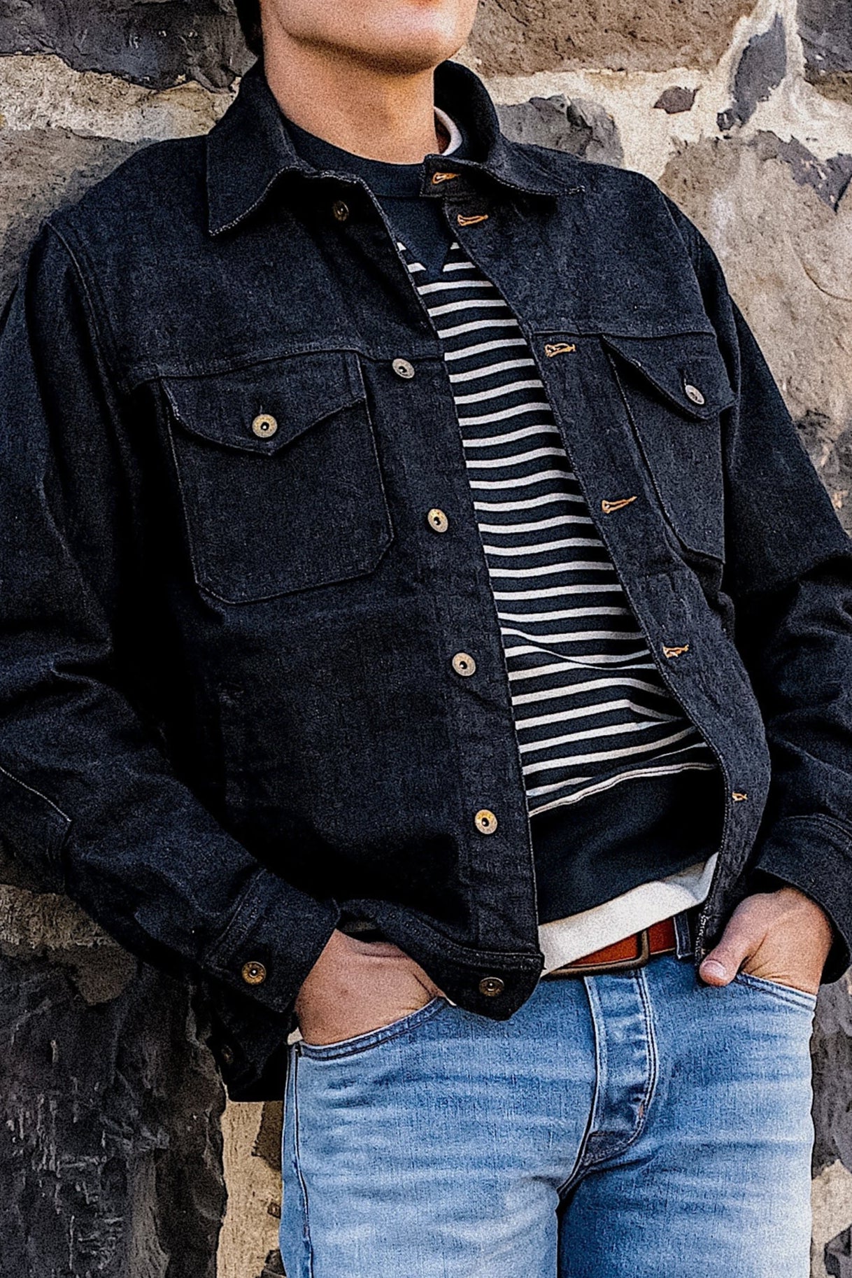 Black denim jacket with button closures and chest pockets, layered over a striped sweater and paired with blue jeans.