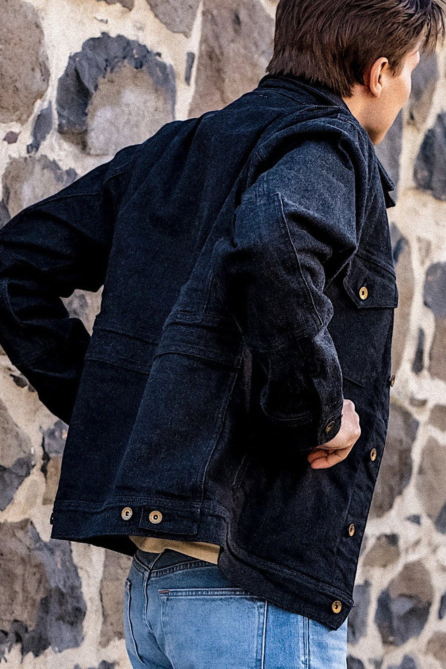 Dark denim jacket with button closures and chest pockets, worn over a beige shirt and blue jeans against a stone wall background.