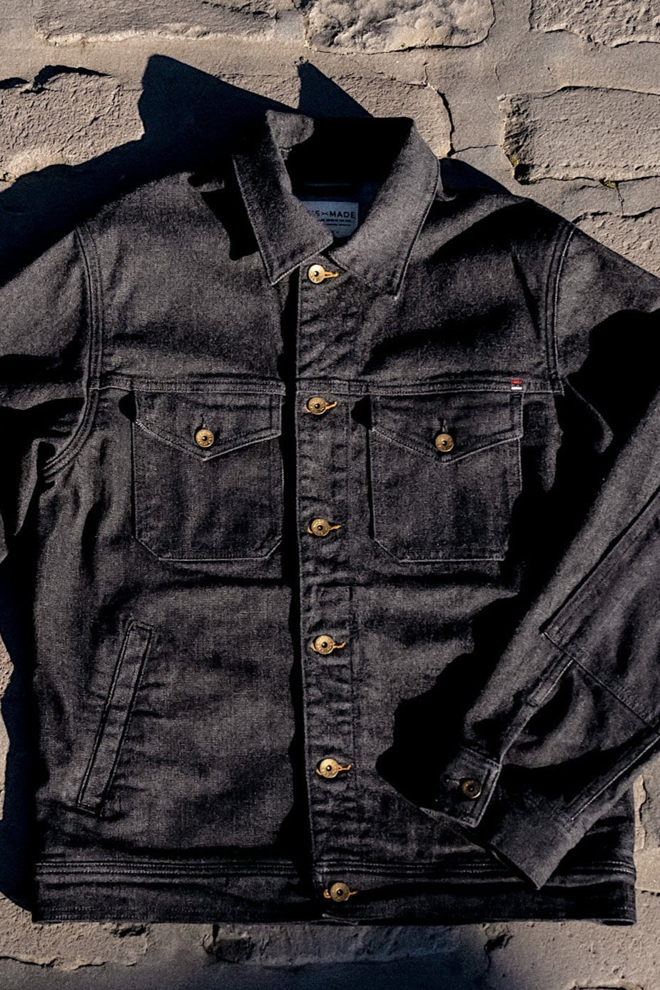 Denim jacket in black with button closures and chest pockets, laid flat on a textured stone surface in sunlight.