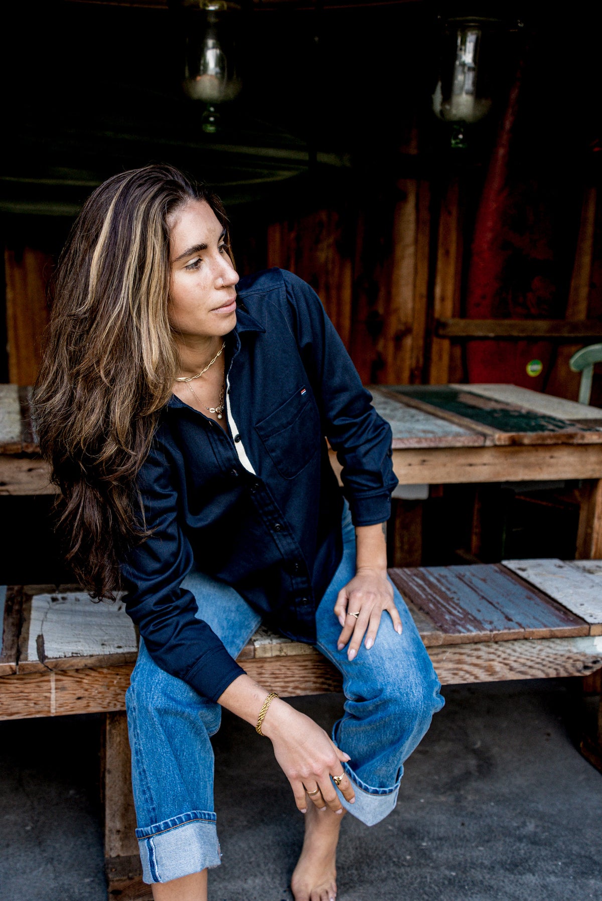 Woman in a navy button-up work shirt and jeans seated in a rustic workshop.