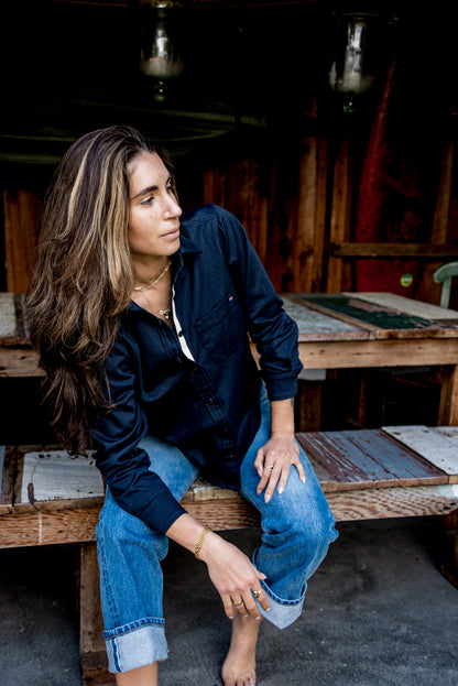 Woman in a navy button-up work shirt and jeans seated in a rustic workshop.