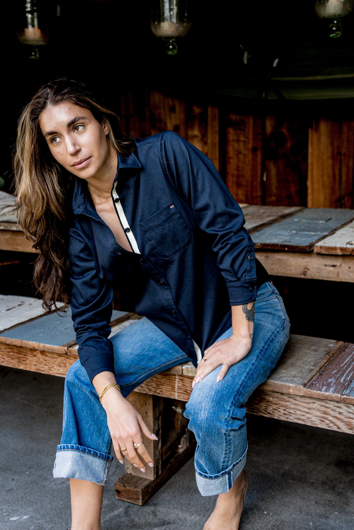 Woman seated on a wooden bench wearing a navy cotton work shirt with cuffed blue jeans.