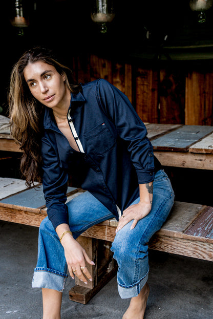 Woman seated on a wooden bench wearing a navy cotton work shirt with cuffed blue jeans.