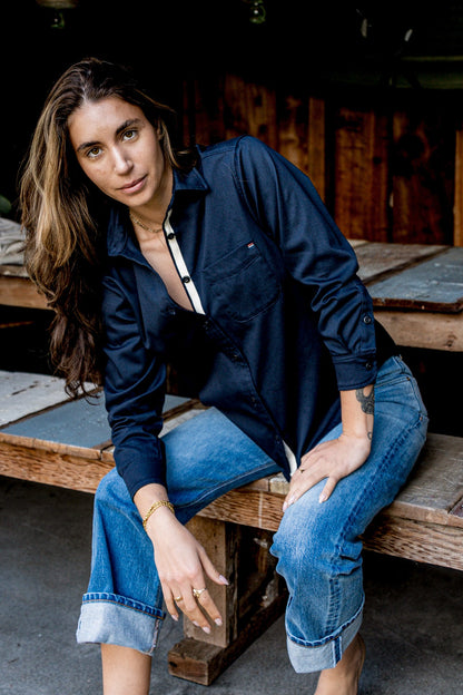 Woman leaning on a workbench in an American made navy cotton shirt and cuffed jeans.