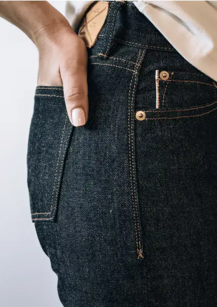 idney Fit Jean in Raw Selvedge Denim color, featuring high-rise and straight-leg styling, worn by a woman with a tucked-in white shirt. The focus is on the back pocket and stitching details against a clean, neutral white background