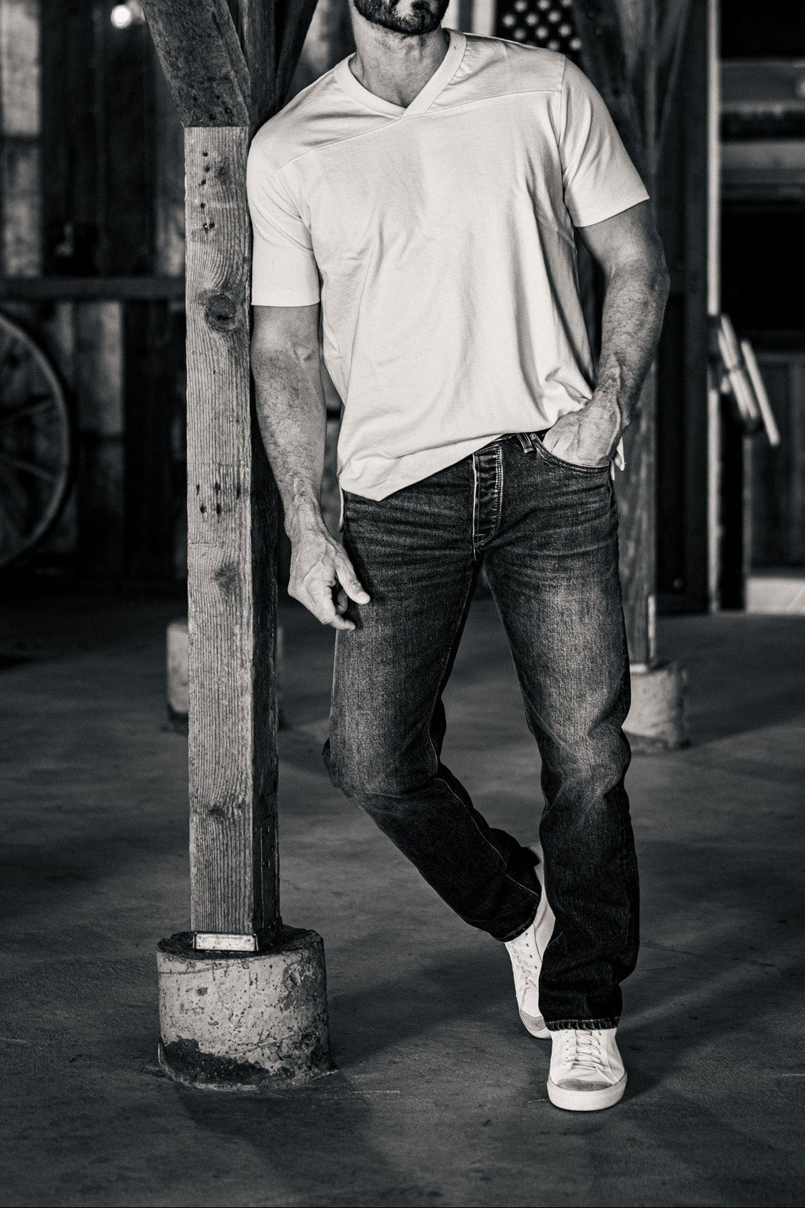 Black-and-white photo of a man wearing men's straight-leg denim jeans and a white v-neck t-shirt, leaning against a wooden post in a rustic barn.