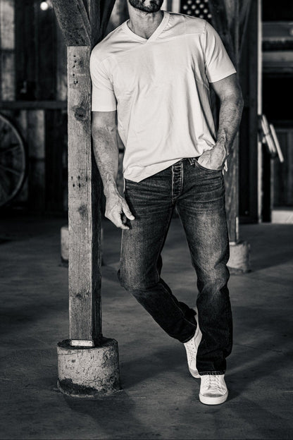 Black-and-white photo of a man wearing men's straight-leg denim jeans and a white v-neck t-shirt, leaning against a wooden post in a rustic barn.