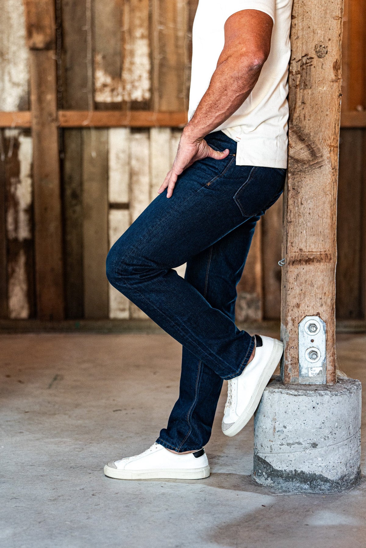 Man leaning against a rustic wooden pillar in an industrial warehouse, wearing men's dark wash slim straight jeans, a white t-shirt, and white slip-on sneakers.