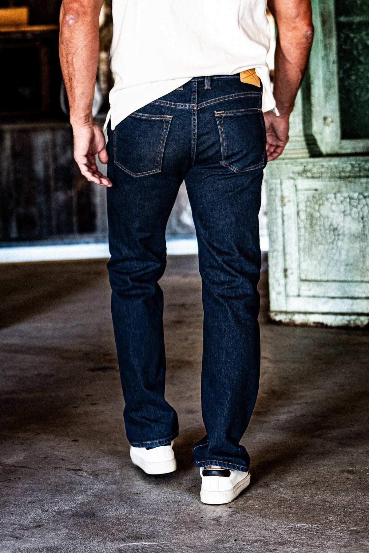 Back view of a man in men's dark wash slim straight jeans, a white t-shirt, and white sneakers, standing in an industrial space with a green door.