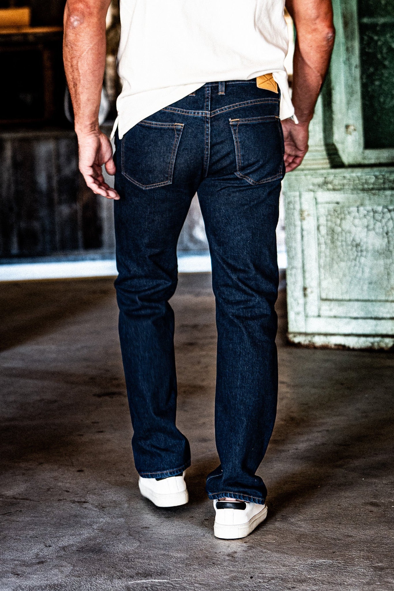 Back view of a man in men's dark wash slim straight jeans, a white t-shirt, and white sneakers, standing in an industrial space with a green door.