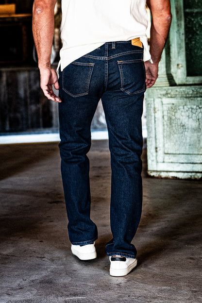 Back view of a man in men's dark wash slim straight jeans, a white t-shirt, and white sneakers, standing in an industrial space with a green door.