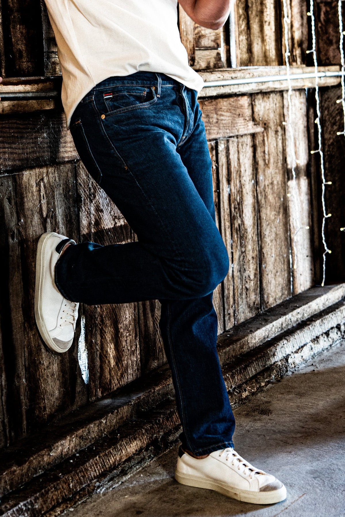 Man leaning against a rustic wooden wall wearing men's dark wash slim straight jeans and a white t-shirt.