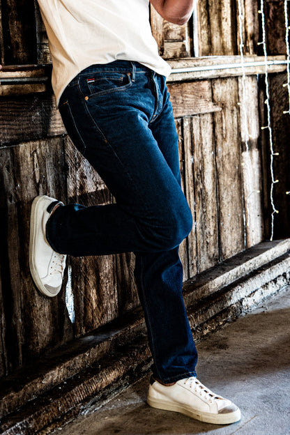 Man leaning against a rustic wooden wall wearing men's dark wash slim straight jeans and a white t-shirt.