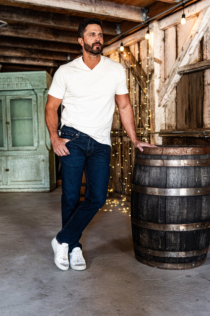 Man standing in a rustic barn wearing men's dark wash slim straight jeans, a white polo shirt, and white sneakers, next to a wooden barrel with string lights.