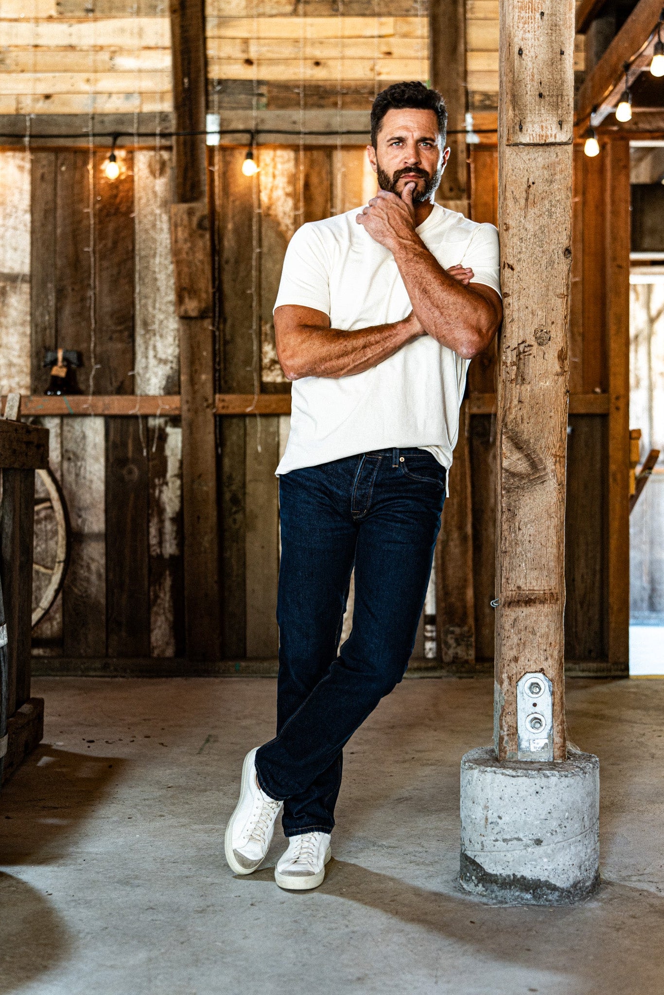 Bearded man leaning against a rustic wooden pillar in a barn, wearing men's dark wash slim straight jeans, a white polo shirt, and white sneakers.