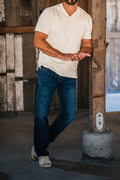Bearded man wearing men's medium wash straight jeans and a cream v-neck t-shirt, leaning against a wooden post in a rustic barn.