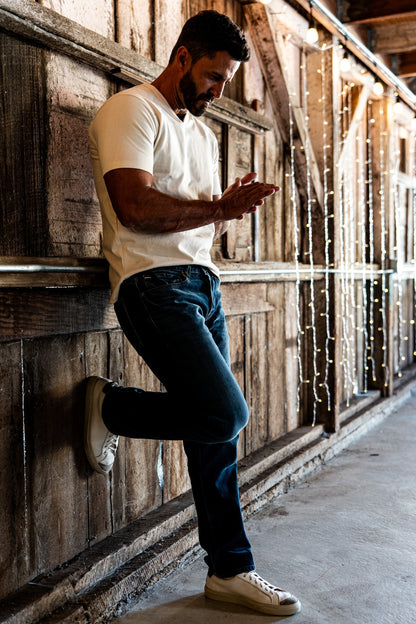 Man leaning against a rustic wooden wall wearing men's medium wash slim straight jeans, a white crewneck t-shirt, and off-white sneakers, with string lights in the background.
