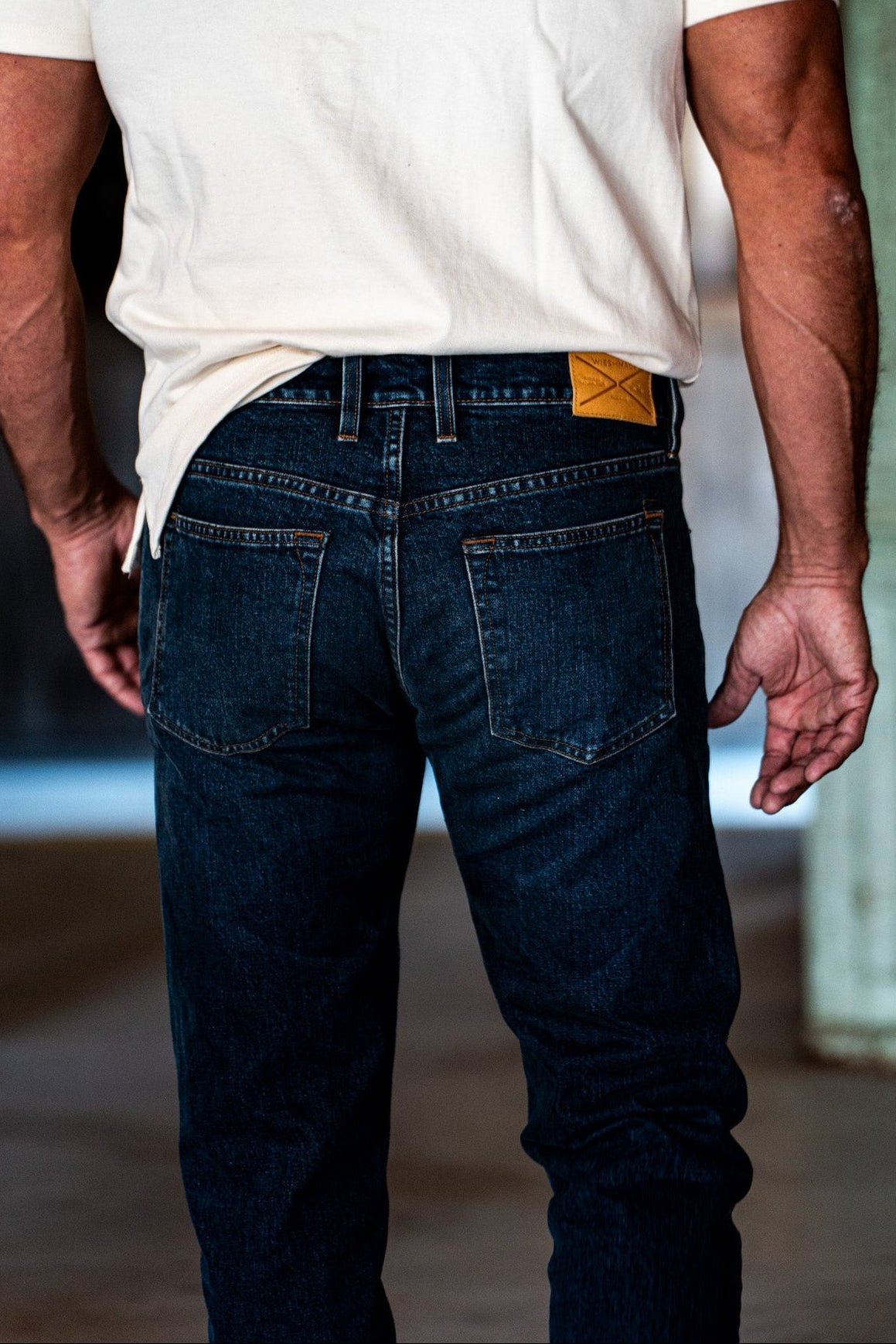 Close-up rear view of men's dark wash denim jeans showing back pocket details and leather brand patch, worn with a white t-shirt.