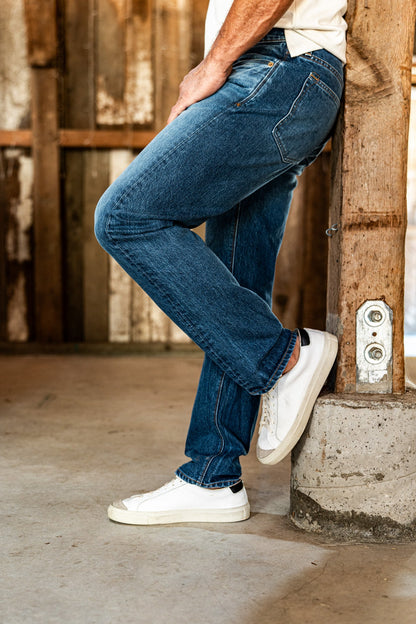 Side view of a man's legs in light wash American made jeans and white sneakers, one knee bent against a rustic wooden post.