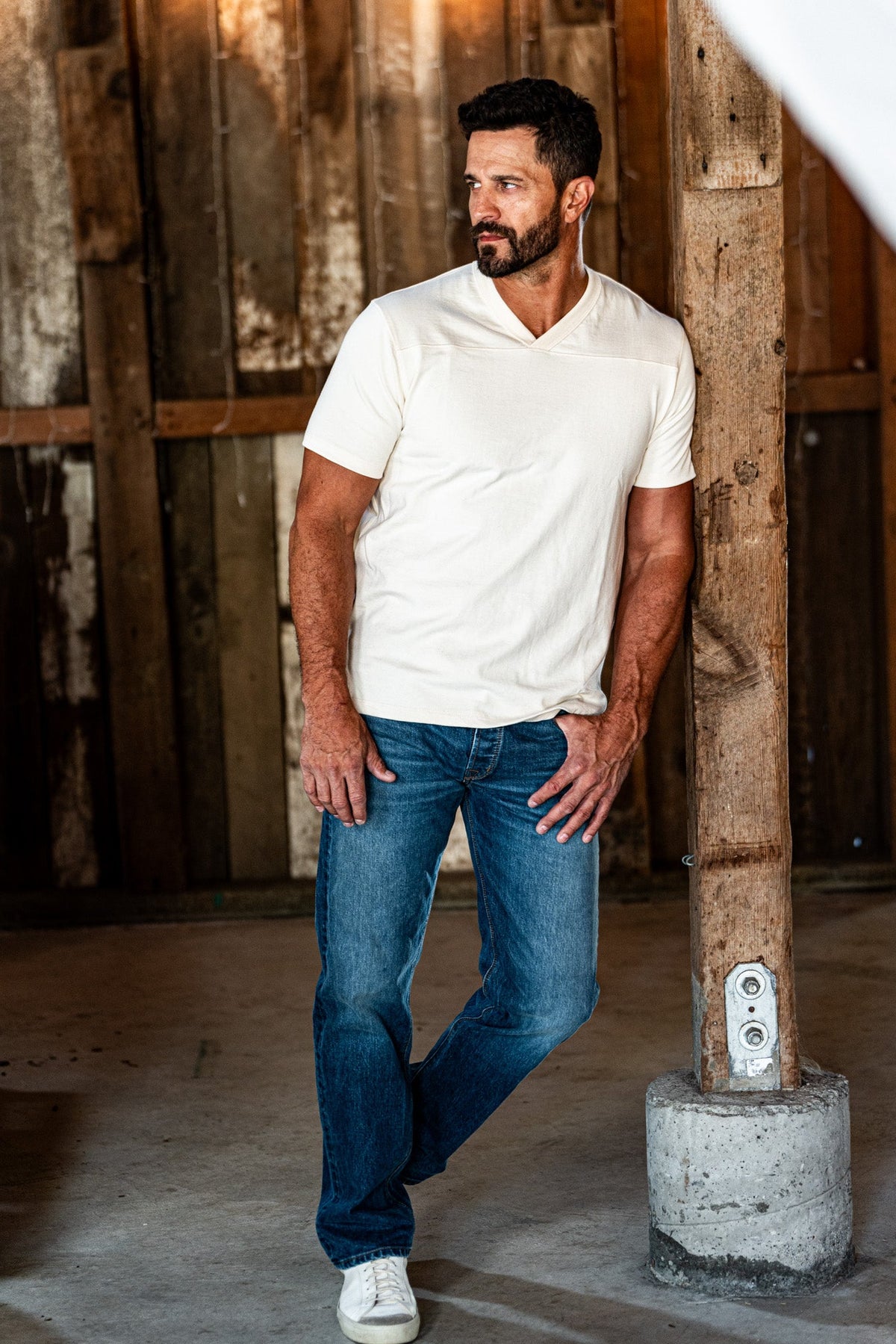 Bearded man in a cream v-neck t-shirt and light wash American made jeans leaning against a wooden beam in a rustic barn.