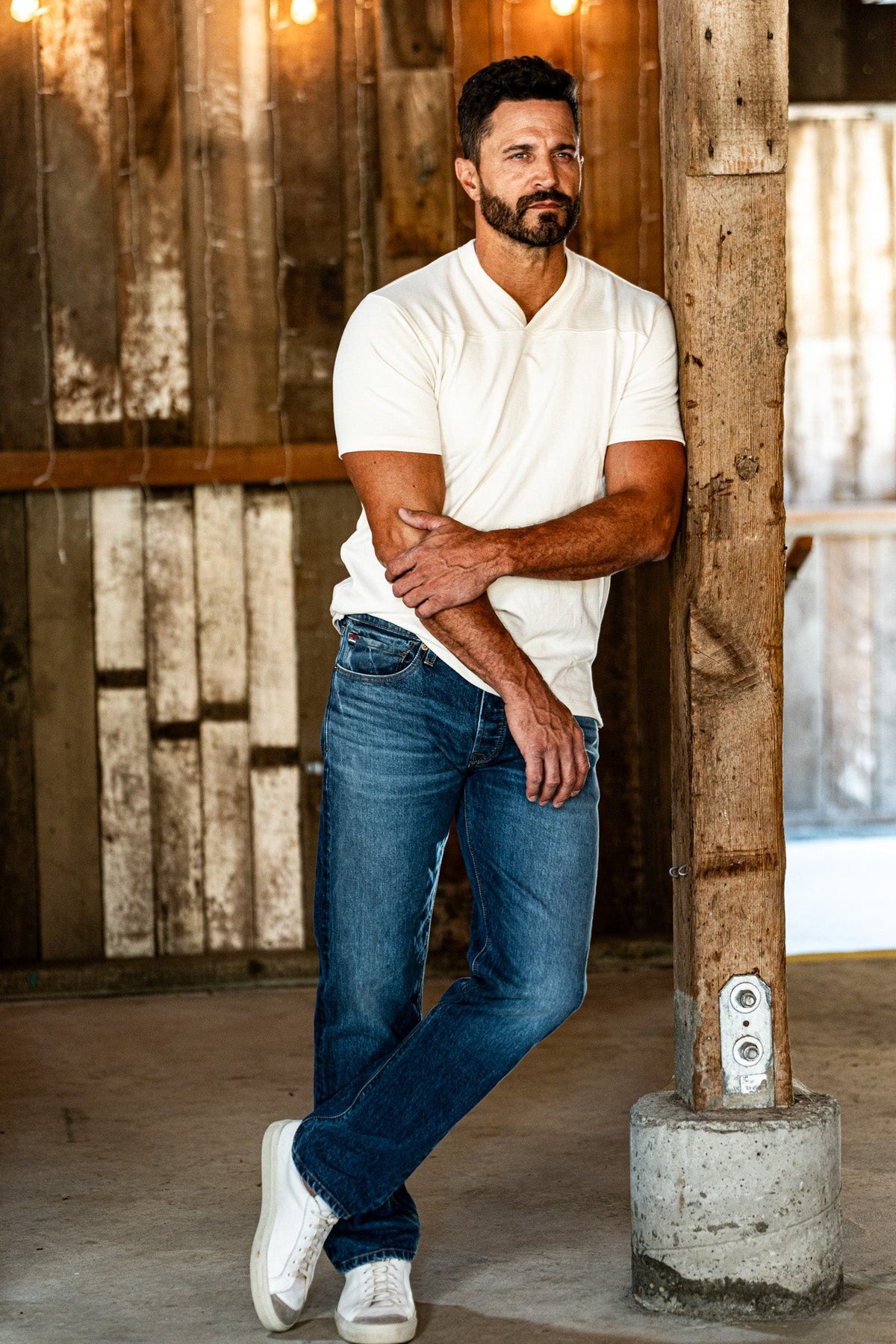 Bearded man in a cream knit shirt and light wash American made jeans leaning against a barn post under string lights.