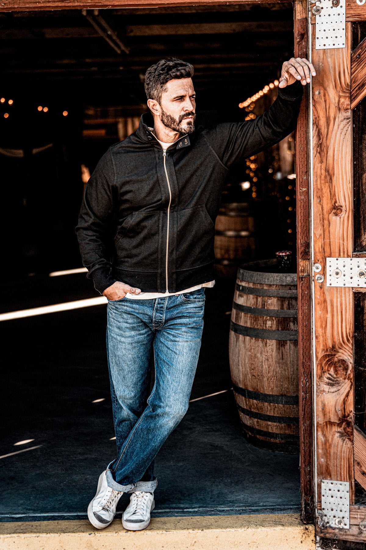 Man in a men's charcoal zip-up moto jacket and jeans standing in a rustic barn doorway near a wine barrel