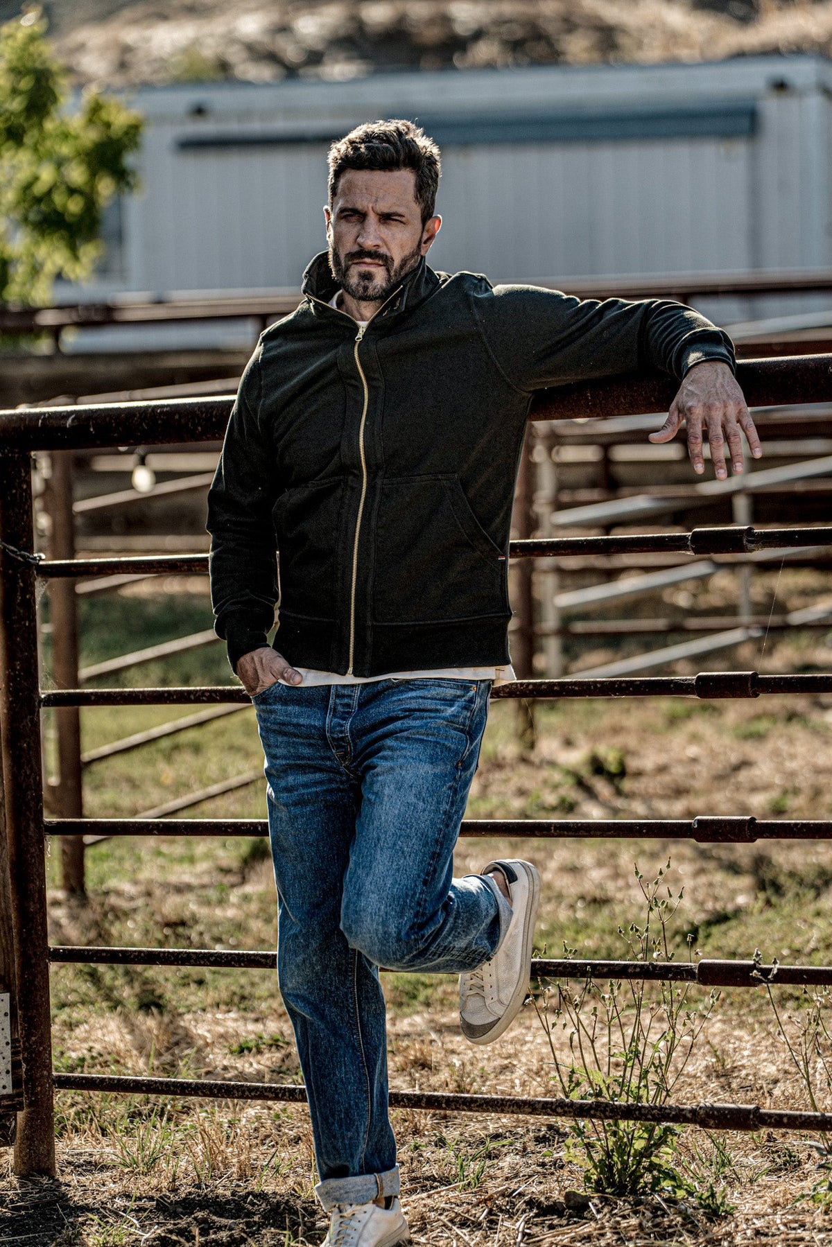 Man wearing a men's charcoal zip-up moto jacket and jeans leaning against a metal farm gate