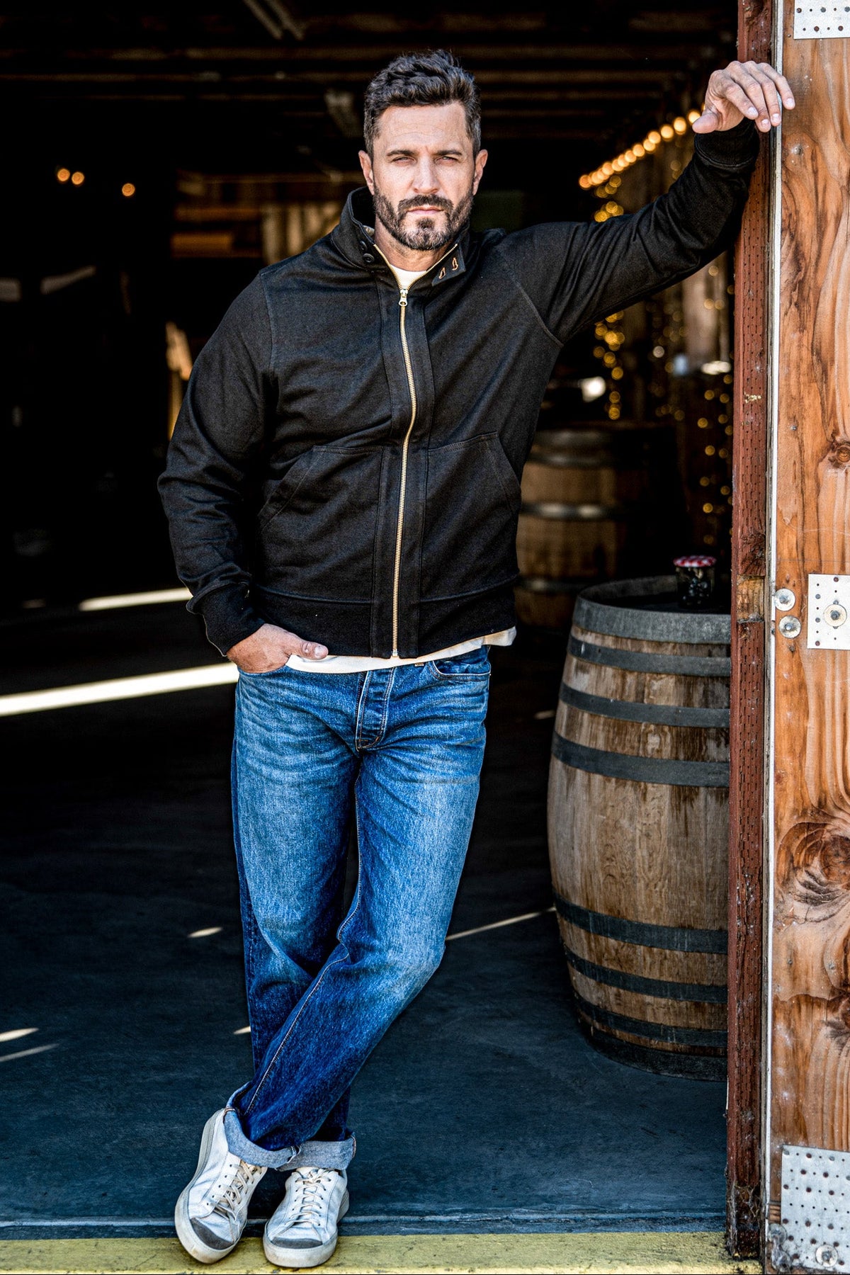 Man wearing a men's charcoal zip-up moto jacket and cuffed jeans posing in a wine cellar doorway