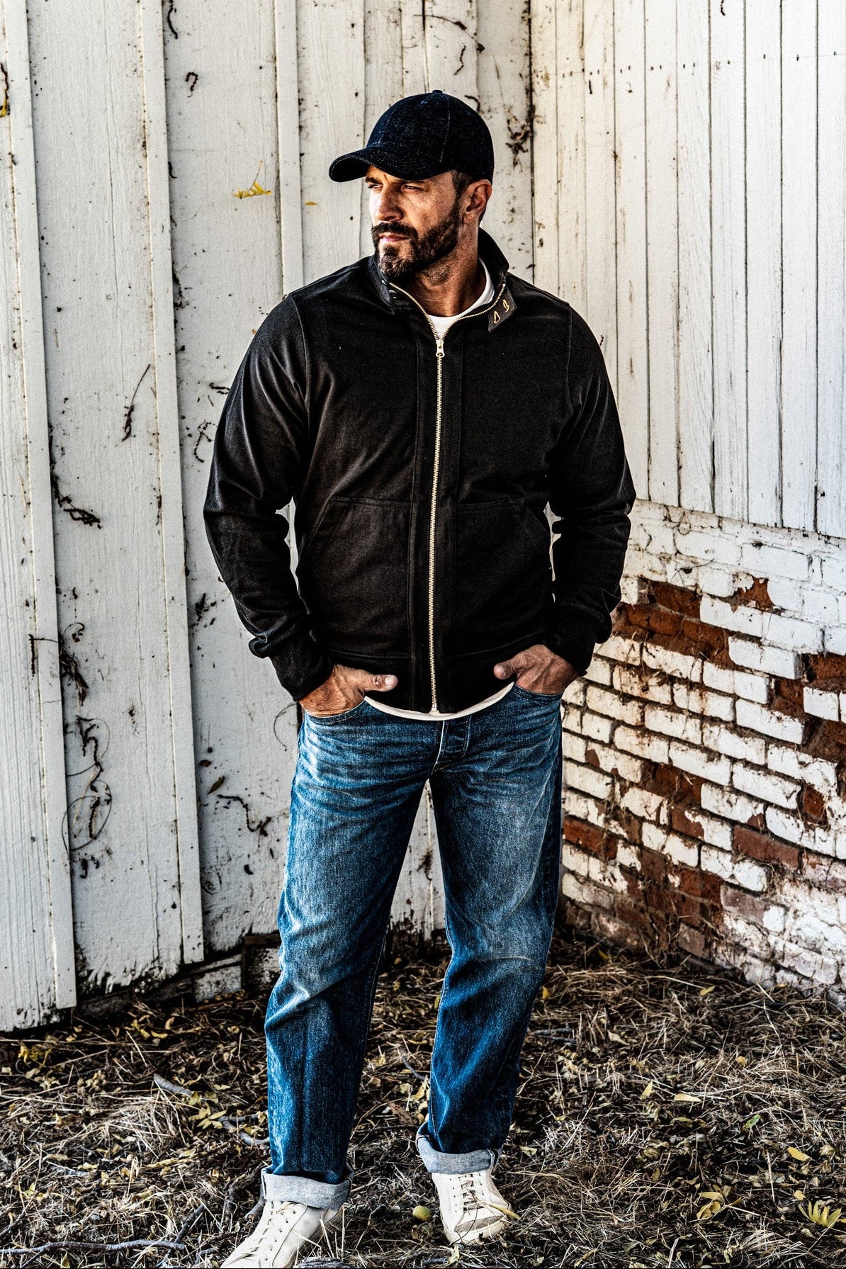 Man wearing a men's charcoal zip-up moto jacket with a baseball cap and cuffed jeans, standing against a rustic white wall