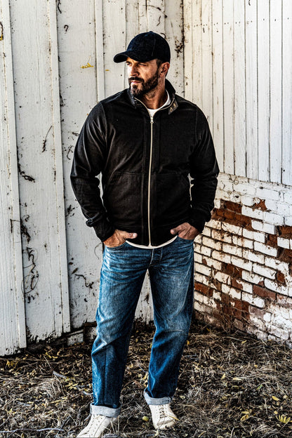 Man wearing a men's charcoal zip-up moto jacket with a baseball cap and cuffed jeans, standing against a rustic white wall