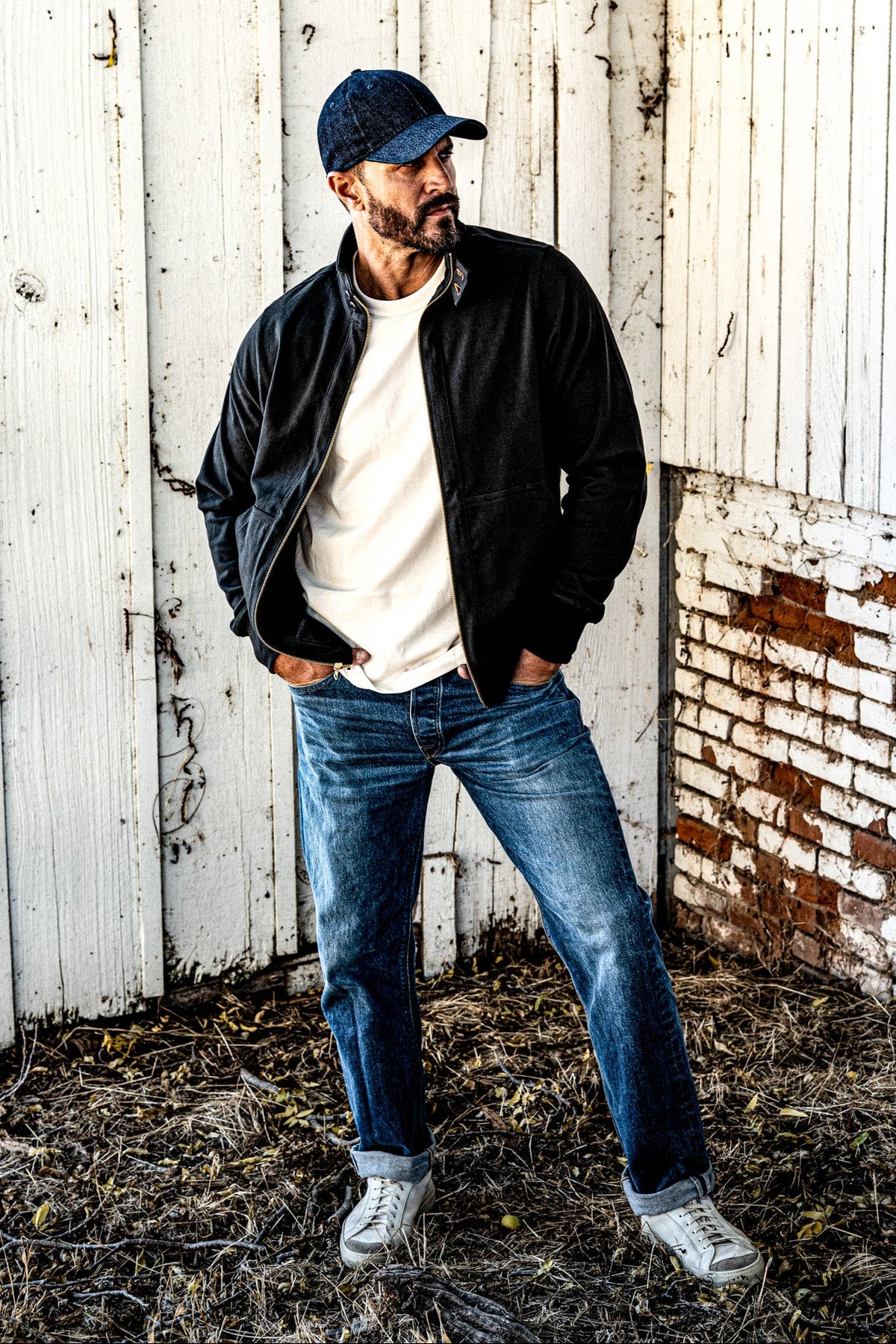 Man in a men's charcoal zip-up moto jacket, baseball cap, and jeans standing against a weathered white wall