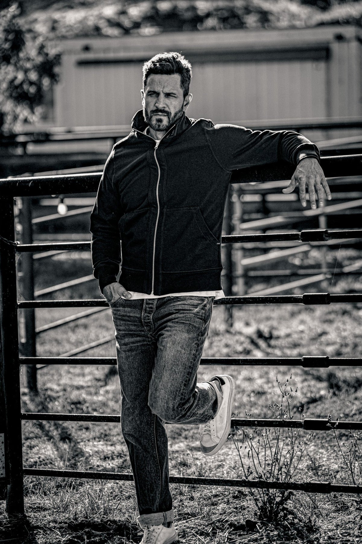 Black and white photo of a man in a men's zip-up moto jacket and jeans leaning against a farm gate