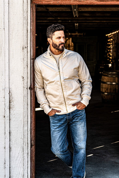 Man wearing a men's cream zip-up moto jacket and jeans standing in a rustic barn doorway