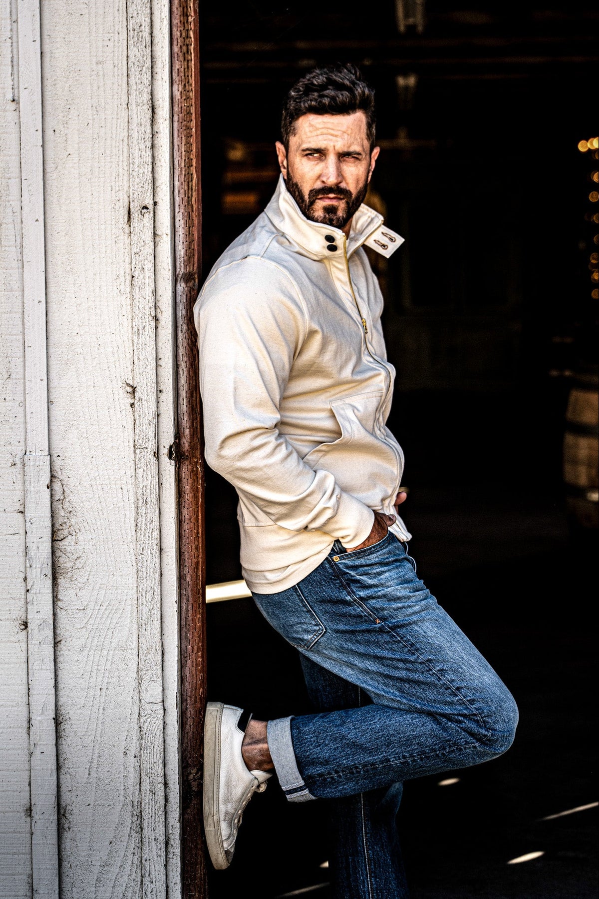 Man wearing a men's cream zip-up moto jacket leaning in a barn doorway, side profile view