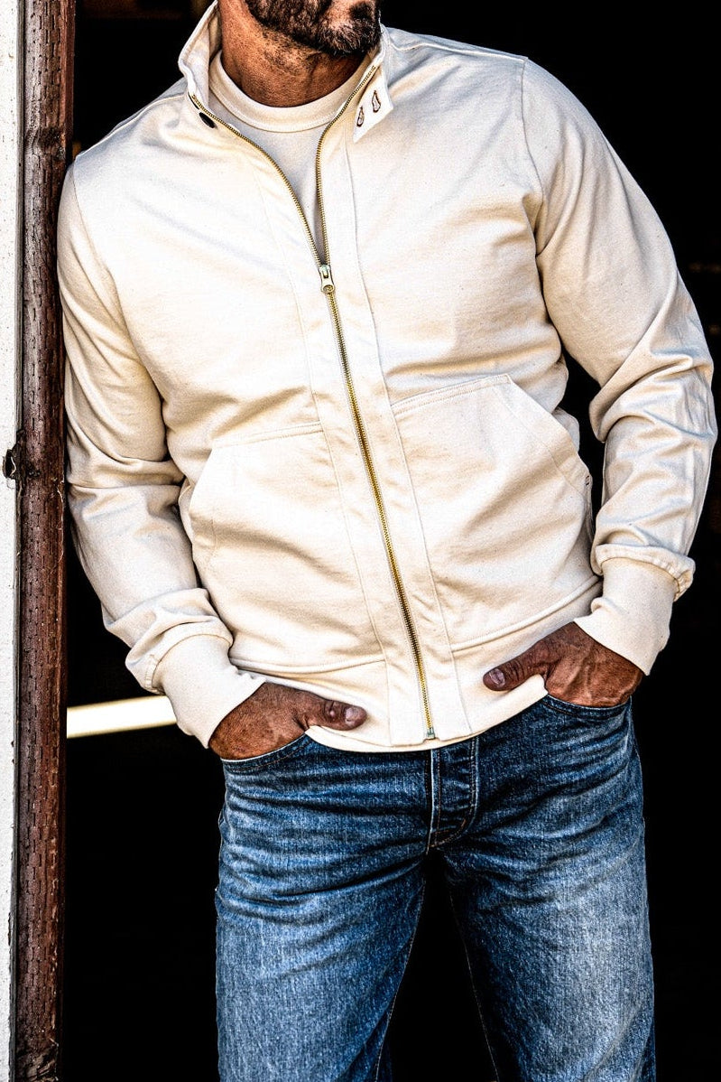 Man in a men's cream zip-up moto jacket and jeans standing beside a wooden post with hands in pockets