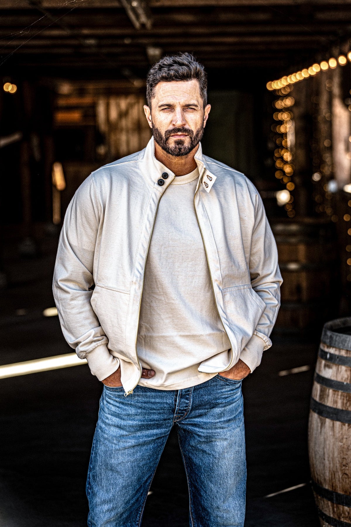 Man wearing an open men's zip-up moto jacket over a tee with jeans beside a wine barrel