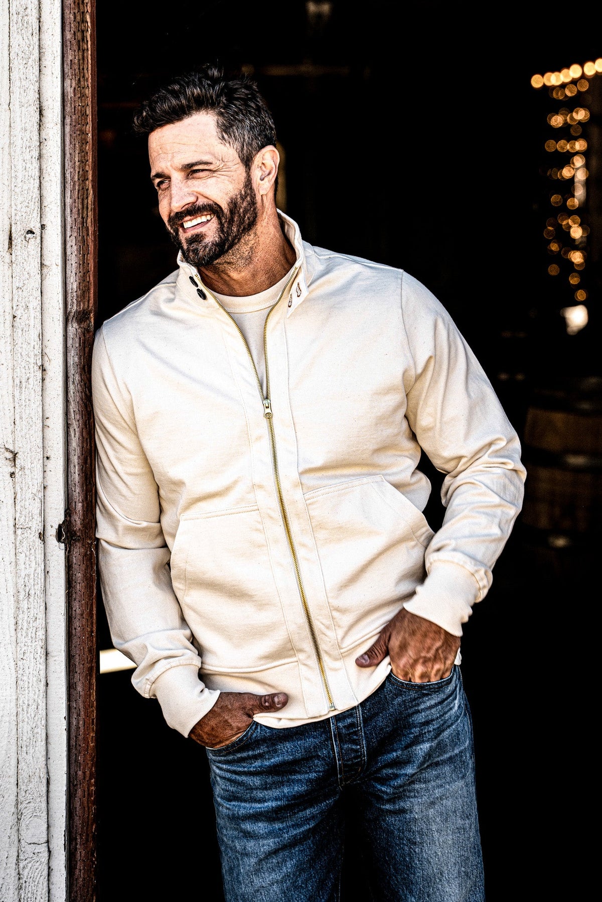Smiling man in a men's zip-up moto jacket and jeans leaning in a barn doorway