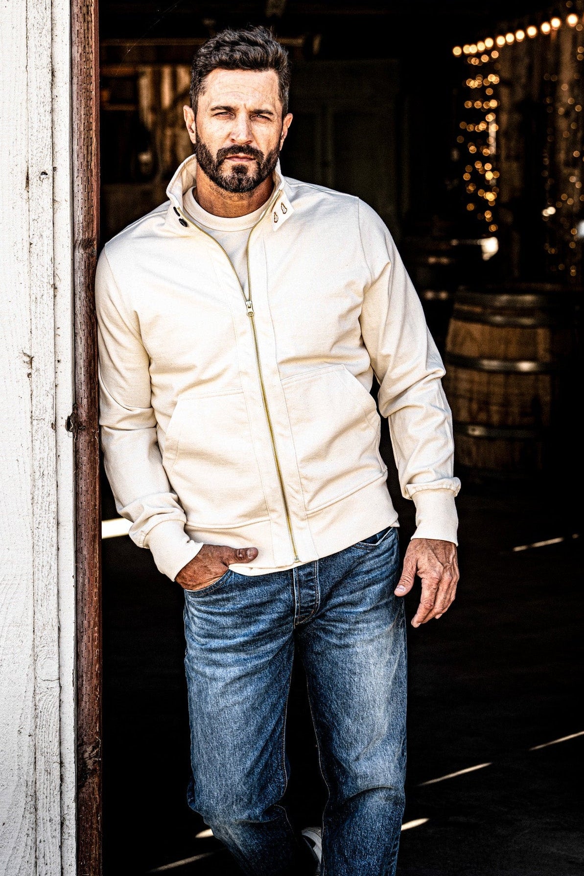 Man wearing a men's cream zip-up moto jacket and jeans standing in a wine cellar doorway
